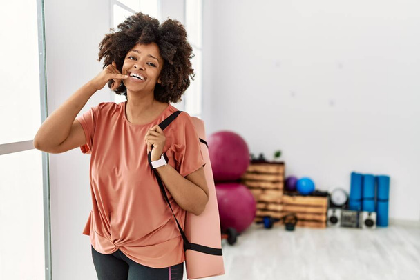 Afro saçlı, pilates odasında yoga minderi tutan, telefonla el ve parmaklarıyla telefonla konuşan Afro-Amerikalı bir kadın. iletişim kavramları.  - Fotoğraf, Görsel