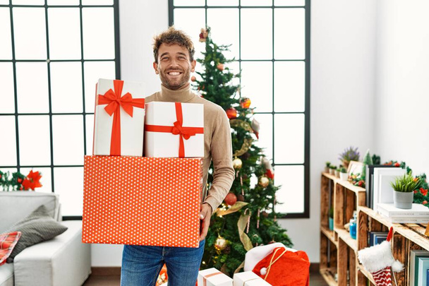 Junger hispanischer Mann lächelt zuversichtlich und hält Weihnachtsgeschenke zu Hause - Foto, Bild