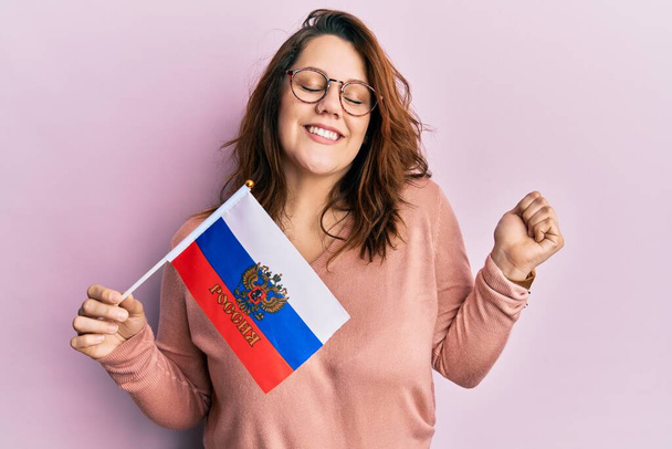 Młoda kaukaska kobieta trzyma rosyjską flagę federacji krzycząc dumny, świętując zwycięstwo i sukces bardzo podekscytowany z podniesionym ramieniem  - Zdjęcie, obraz
