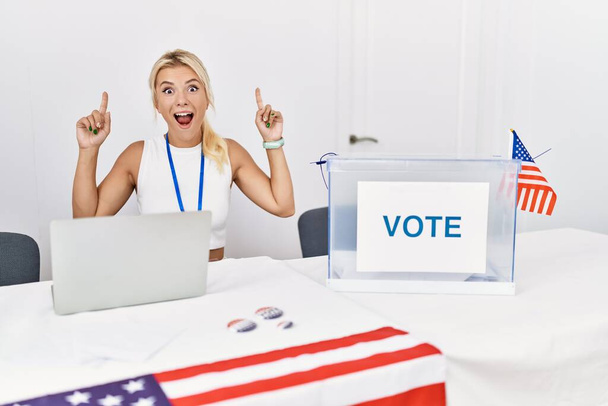 Jeune femme caucasienne à l'élection américaine campagne politique sourire étonné et surpris et pointant vers le haut avec les doigts et les bras levés.  - Photo, image
