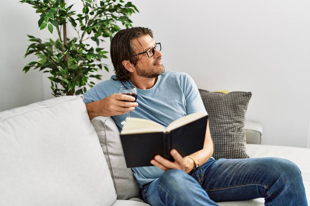 Middle age caucasian man reading book drinking wine at home - Foto, Imagen
