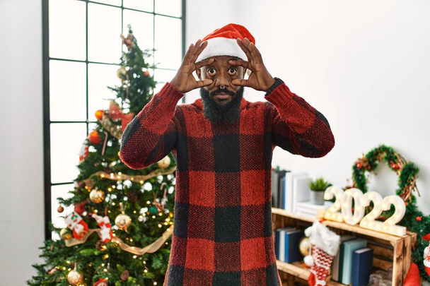 Αφροαμερικάνος που φοράει καπέλο Σάντα Κλάους στέκεται δίπλα στο χριστουγεννιάτικο δέντρο προσπαθώντας να ανοίξει τα μάτια με τα δάχτυλα, νυσταγμένος και κουρασμένος για την πρωινή κούραση  - Φωτογραφία, εικόνα