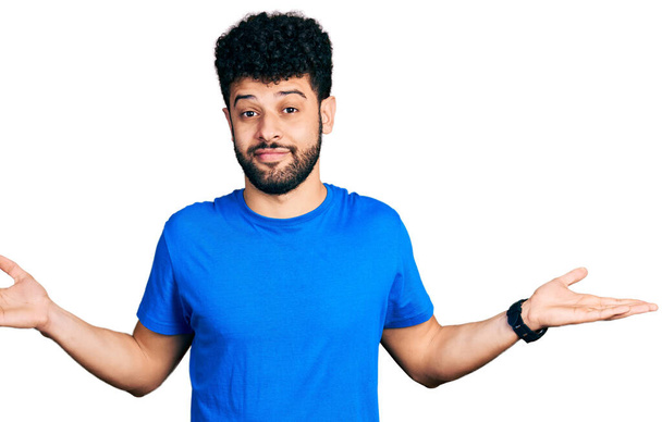Fiatal arab férfi szakállal, alkalmi kék pólóban, tanácstalan és zavaros arckifejezéssel, felemelt karokkal és kezekkel. kétség.  - Fotó, kép