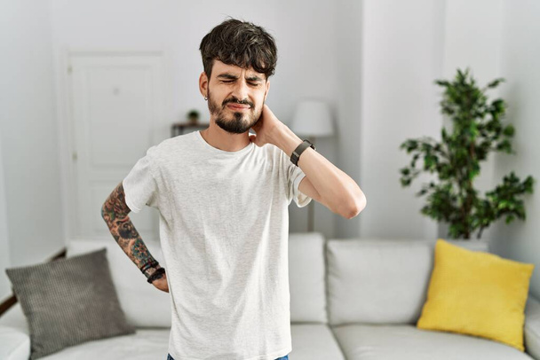 Spaanse man met baard in de woonkamer thuis die last heeft van nekpijn, nek met de hand aanraken, spierpijn  - Foto, afbeelding