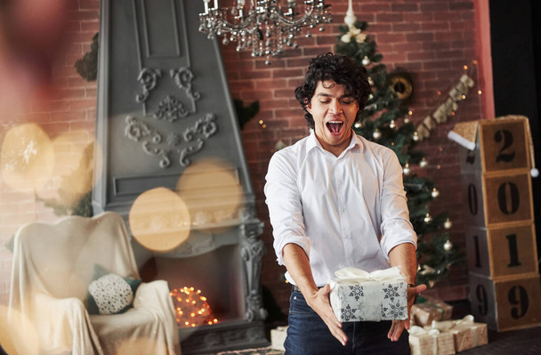 Gifts time. Young man standing in the beautiful decorated room and holding white box with present in the New year time. - Foto, afbeelding