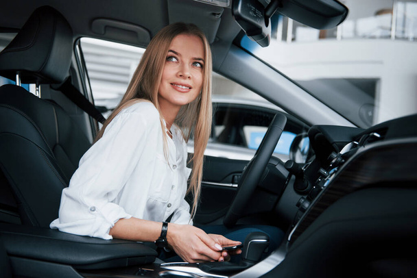 Holds smartphone and smiling. Beautiful blonde girl sitting in the new car with modern black interior. - Foto, Imagem