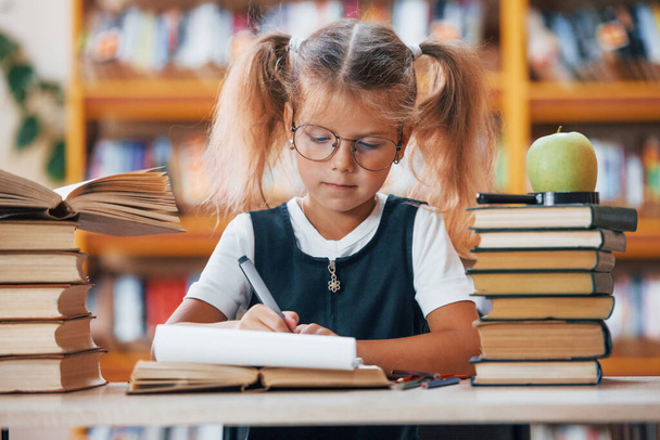 Cute little girl with pigtails is in the library. Apple on the books. - Φωτογραφία, εικόνα