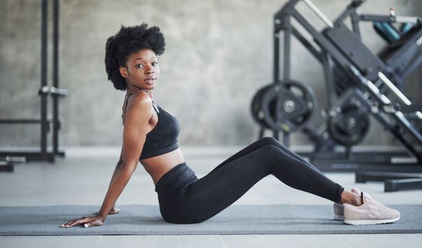 On grey colored karemat. African american woman with curly hair and in sportive clothes have fitness day in the gym. - Photo, image