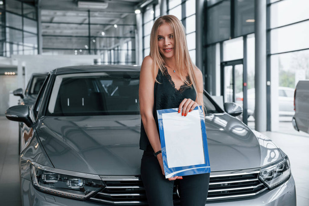 In black pants. Girl and modern car in the salon. At daytime indoors. Buying new vehicle. - Фото, изображение