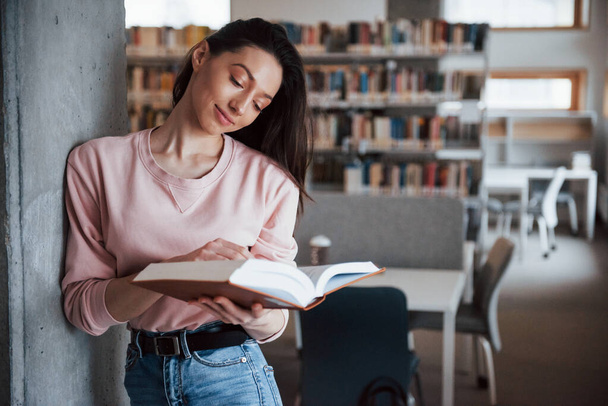 Leaning on the wall. Brunette girl in casual clothes having good time in the library full of books. - Photo, Image