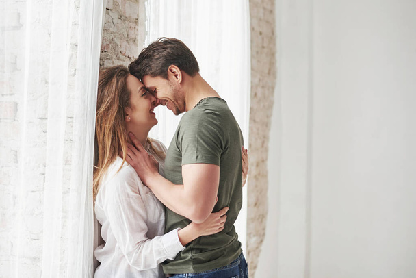 Attractive youth. Lovely couple kissing and enjoying each other near the window of their new home. - Fotoğraf, Görsel