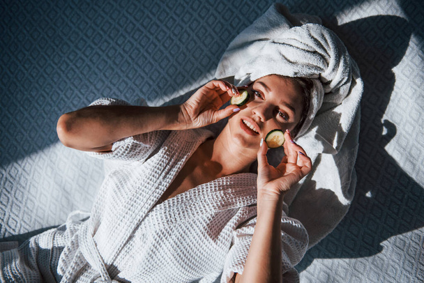 Young positive woman with towel on head lying on the bed with cucumber. - Photo, Image