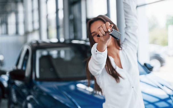 Excited with her new car. Holds keys in hand. Young woman in white official clothes stands in front of blue automobile indoors. - Zdjęcie, obraz