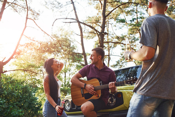 Singing songs together. Friends have nice weekend outdoors near theirs green car with acoustic guitar. - Valokuva, kuva