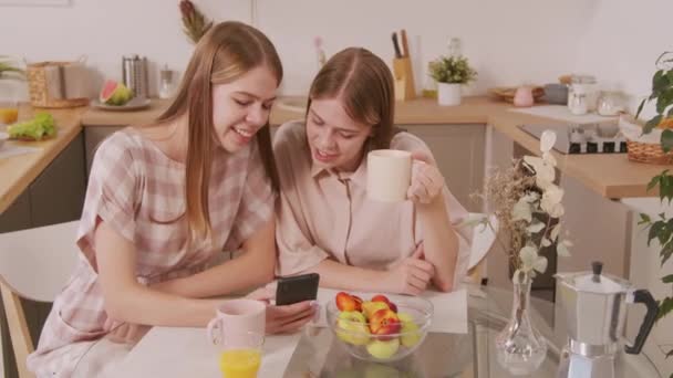 Handheld laukaus onnellinen nuori kaksoissisko istuu pöydässä viihtyisässä keittiössä ja nauraa katsellen matkapuhelin ja chattailuun yli aamulla kahvia - Materiaali, video