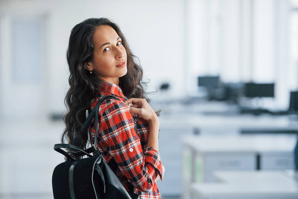 Computer screens behind. Portrait of attractive young woman standing in the office with black bag. - 写真・画像