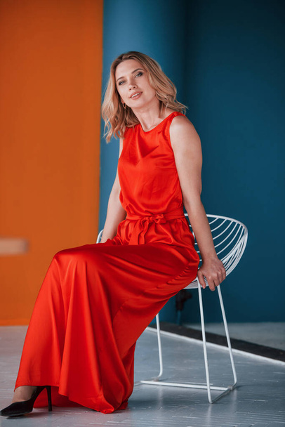 Ready for the party. Beautiful blonde in festive red dress is in the room with orange and blue colored walls and white chair. - Фото, изображение