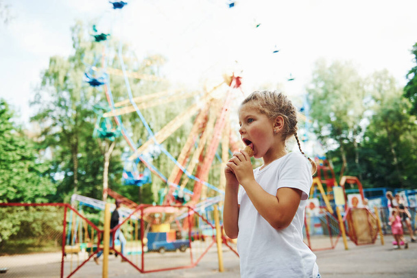 Cute little girl eats ice cream in the park at daytime near attractions. - Foto, Imagen