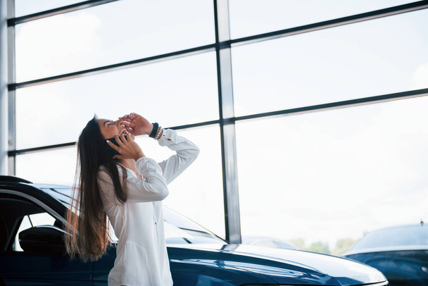 Having conversation by the phone. Young woman in white official clothes stands in front of blue automobile indoors. - Foto, Bild