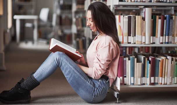 Sitting and reading. Brunette girl in casual clothes having good time in the library full of books. - Photo, image