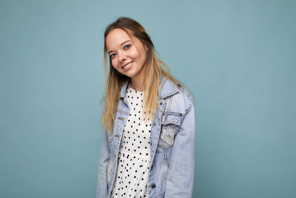 Portrait of young beautiful smiling hipster blonde woman in trendy dress and jean jacket. Sexy carefree female person posing isolated near blue wall in studio. Positive model with natural makeup. Copy - Фото, изображение