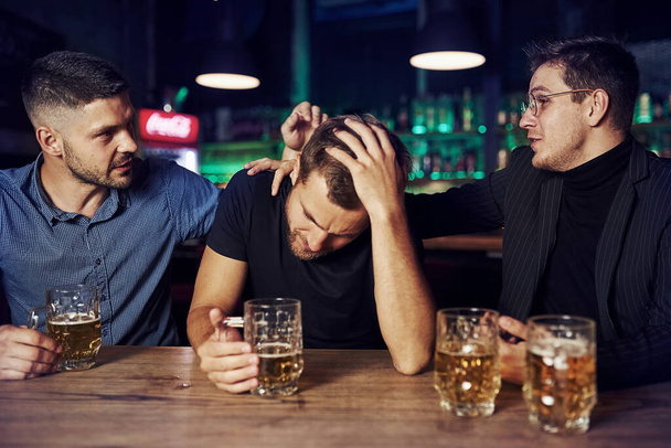 Three male friends in the bar. Supporting sad friend. Unity of people. With beer on the table. - Foto, Imagen