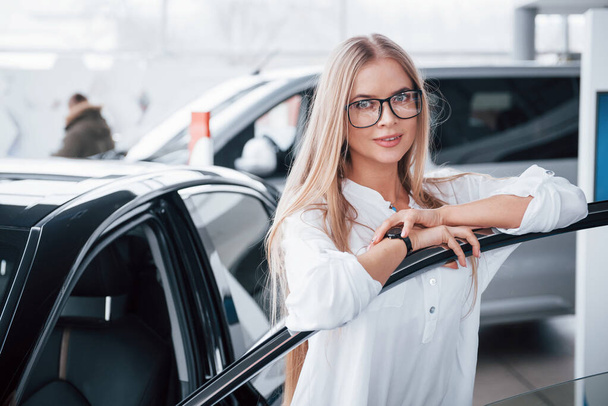 Cute girl in eyewear stands near the car in auto saloon. Probably her next purchase. - Fotografie, Obrázek