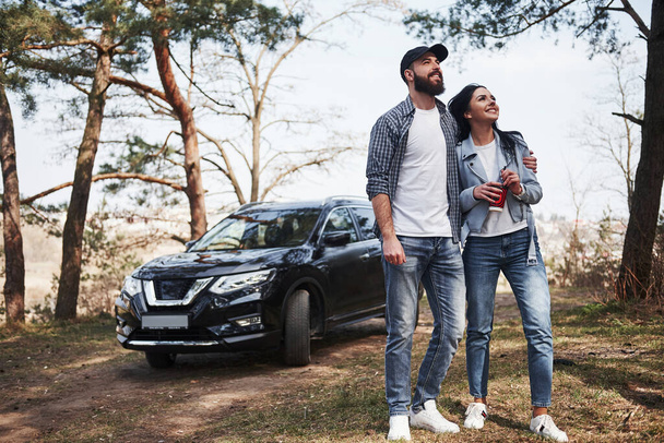 Bearded guy embracing his girlfriend. Embracing and enjoying the nature. Couple have arrived to the forest on their brand new black car. - Foto, imagen