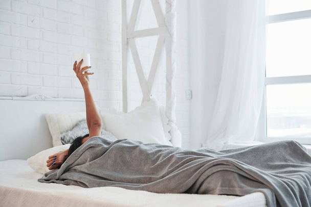Brunette is not going anywhere today. Woman lying under blanket and raising hand up with white cup of coffee in it. - Foto, Bild