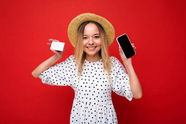 Tiro de bela mulher loira sorridente feliz em roupas de verão mostrando smartphone com tela vazia para simular e cartão de crédito isolado no fundo vermelho. Conceito de pagamento - Foto, Imagem