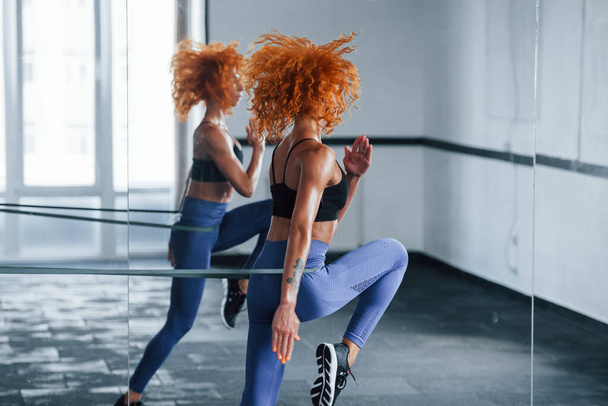 Running near the mirror. Sporty redhead girl have fitness day in gym at daytime. Muscular body type. - Foto, Imagem