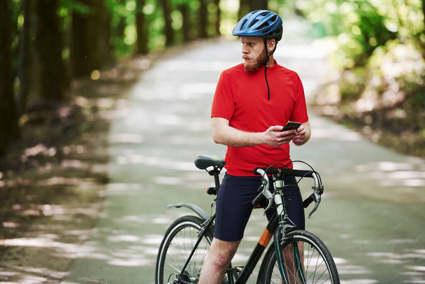 Texting to a friend. Cyclist on a bike is on the asphalt road in the forest at sunny day. - Photo, image