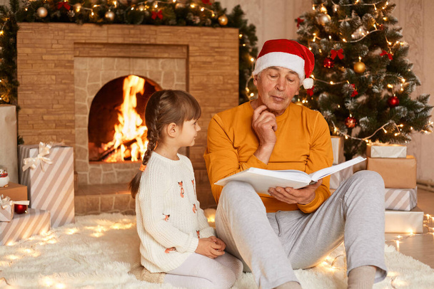 Indoor shot of little granddaughter with pigtails in white pullover and grandfather wearing orange sweater and santa claus hat sitting near Christmas tree and fireplace, reading book together. - Fotografie, Obrázek