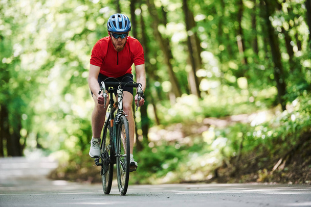 Sportive wear. Cyclist on a bike is on the asphalt road in the forest at sunny day. - Photo, image