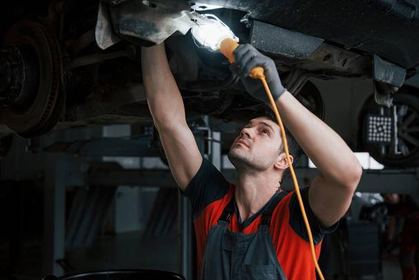 With lantern in hand. Man at the workshop in uniform fixes broken parts of the modern car. - Photo, Image