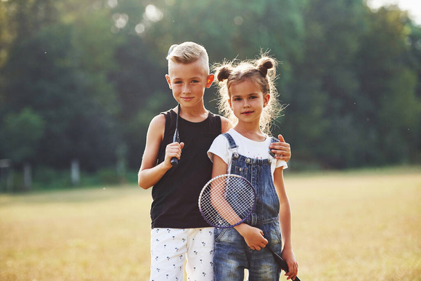 Portrait of boy and girl that standing in the field at sunny day with tennis rackets in hands. - Fotoğraf, Görsel