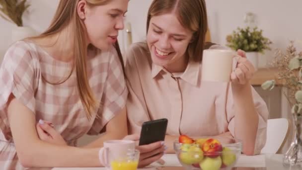 Снимок веселых сестер-близнецов, сидящих за столом на уютной кухне и сидящих, глядя в мобильный телефон, а утром пьющих кофе - Кадры, видео