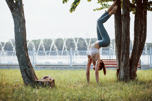 Fountain at background. Young woman with slim type of body does exercises in the park. - Photo, Image