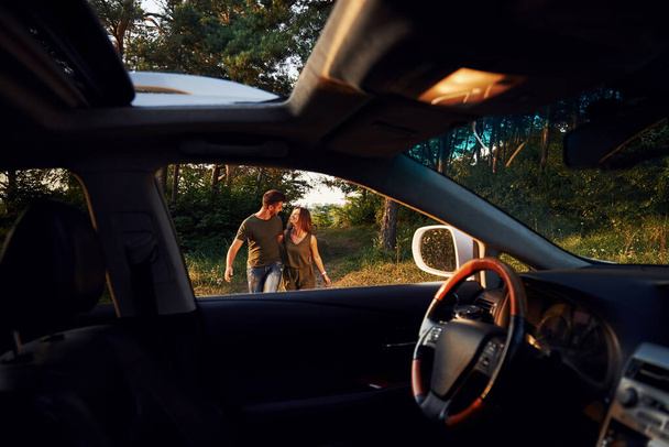 Θέα από το εσωτερικό του αυτοκινήτου. Τιμόνι, πλαϊνός καθρέφτης. Το όμορφο νεαρό ζευγάρι περνάει καλά στο δάσος την ημέρα.. - Φωτογραφία, εικόνα