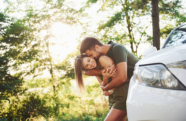 Front part of white car. Beautiful young couple have a good time in the forest at daytime. - Foto, Bild