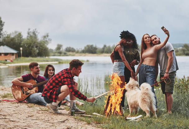 Mann setzt Holz in Brand. Die Gruppe picknickt am Strand. Freunde haben Spaß am Wochenende. - Foto, Bild