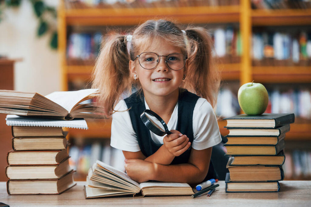 Holds magnifier. Cute little girl with pigtails is in the library. Apple on the books. - Foto, imagen