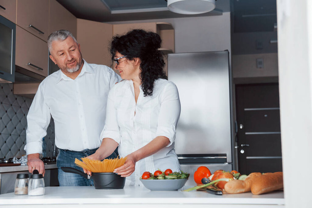Old but happy. Man and his wife in white shirt preparing food on the kitchen using vegetables. - Photo, image