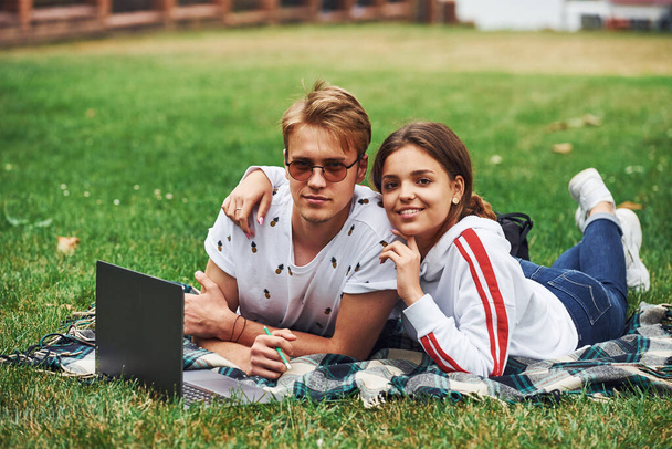 Young couple sits on green grass on the field with their laptop. - Fotó, kép