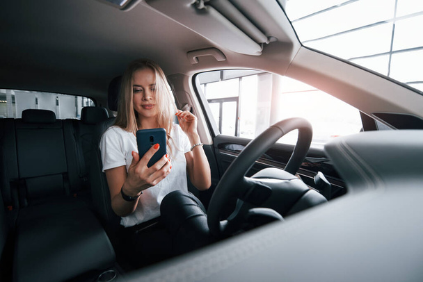 Using blue colored smartphone. Girl in modern car in the salon. At daytime indoors. Buying new vehicle. - Fotografie, Obrázek