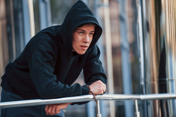 With hood on head. Portrait of young main in black clothes that leaning on the railings and taking a break. - Foto, Bild