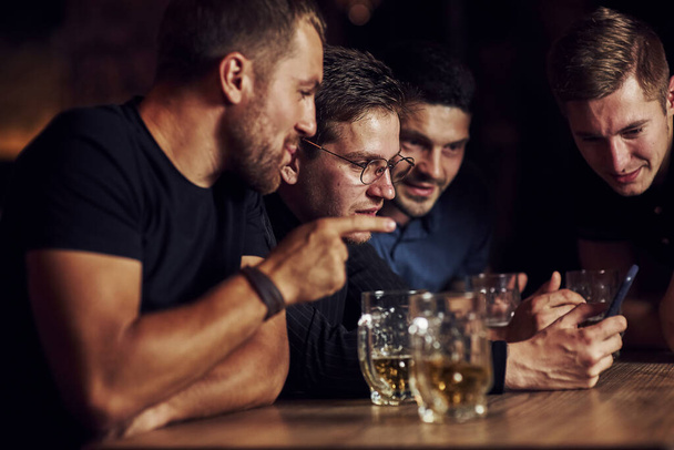Funny content on smartphone. Three friends resting in the pub with beer in hands. Having conversation. - Fotoğraf, Görsel
