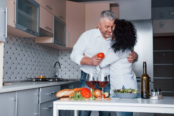 Lovely family couple. Man and his wife in white shirt preparing food on the kitchen using vegetables. - Zdjęcie, obraz