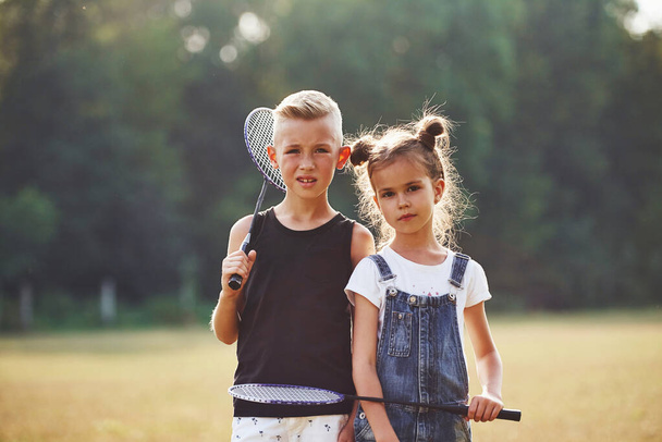 Portrait of boy and girl that standing in the field at sunny day with tennis rackets in hands. - Foto, Imagem