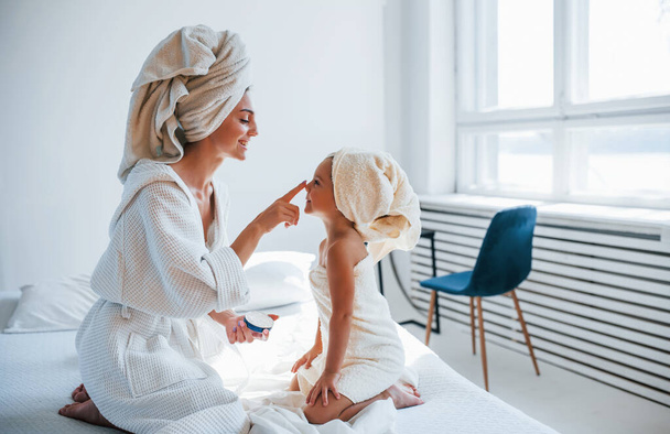 Usando la crema per pulire la pelle. Giovane madre con la figlia hanno giornata di bellezza al chiuso in camera bianca. - Foto, immagini